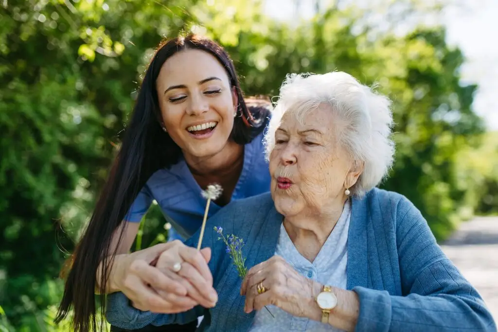 An older woman engages in healthy aging by blowing on a dandelion, while a younger woman smiles beside her, sharing a joyful outdoor moment.