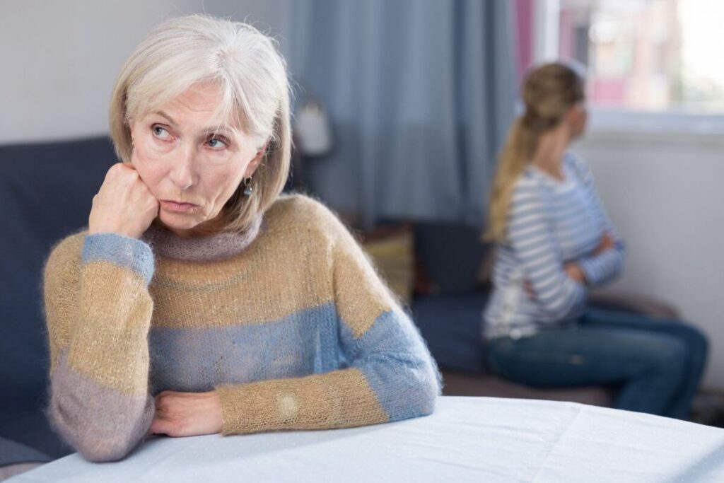 An older woman sits alone at a table looking away with a troubled expression, while a younger woman sits turned away in the background, illustrating the emotional tension that can come with resistance to memory care.