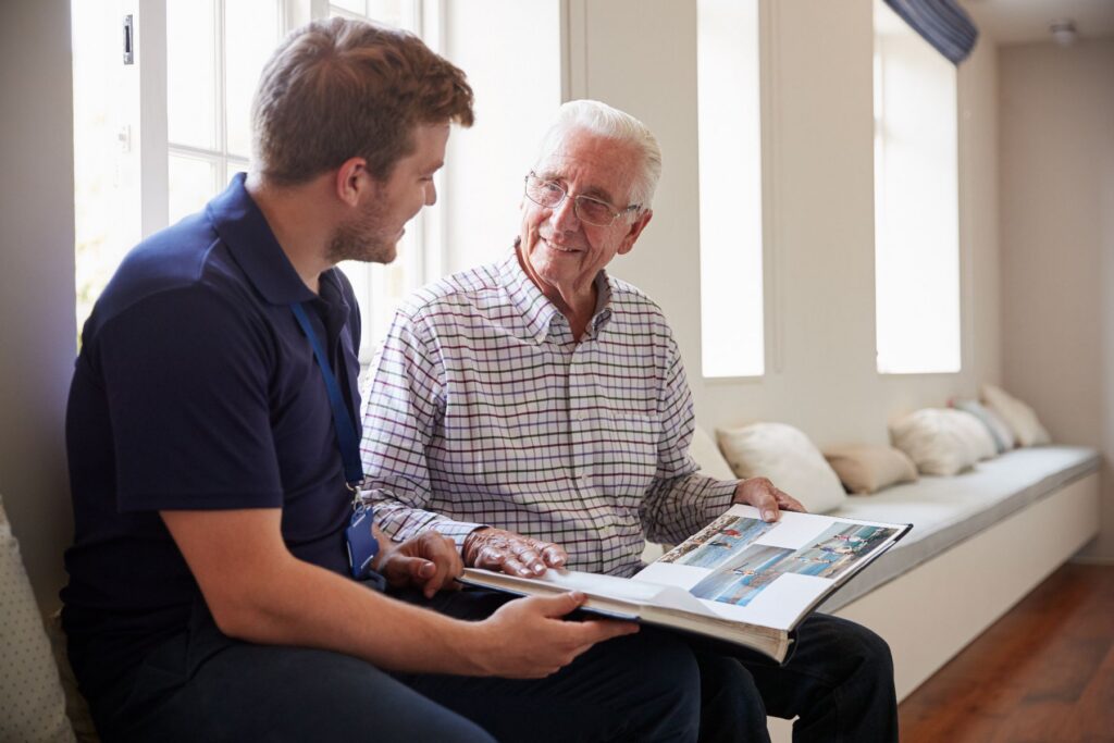 A younger man and an older adult sit together by a sunny window, flipping through a photo album and sharing stories, reflecting the personalized, relationship-centered care that sets assisted living vs. memory care apart in meaningful ways.