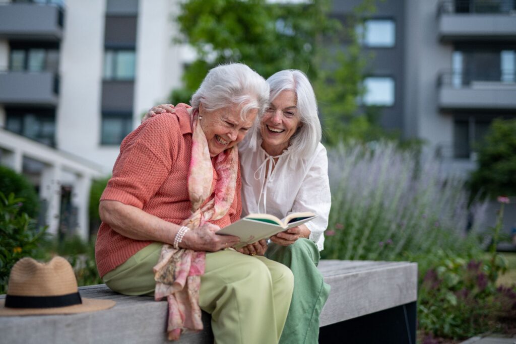 Two older women sit close together on an outdoor bench, laughing warmly as they read from a book, one with her arm around the other's shoulder — a joyful, connected moment that stands in contrast to the subtle early signs of cognitive decline families are often encouraged to watch for.